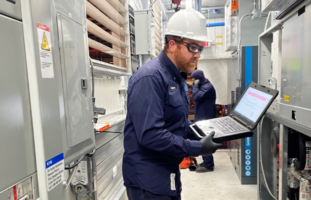 A man in a hard hat and safety glasses operates a laptop, demonstrating safety and technology in a work environment.